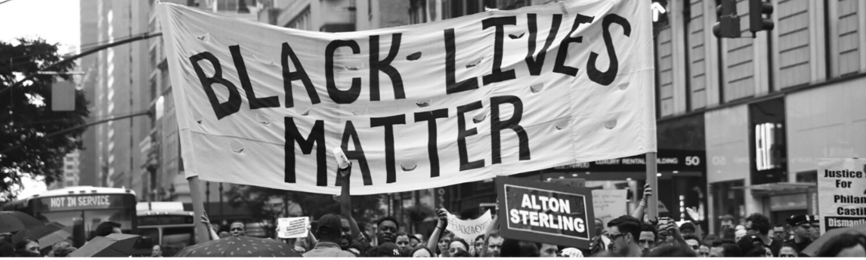 Black and white image of a group of people in a city, peacefully marching holding a sign 'Black Lives Matter'